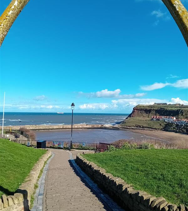 looking down a path to the sea and harbour in front. The sky is blue with clouds in the distance.