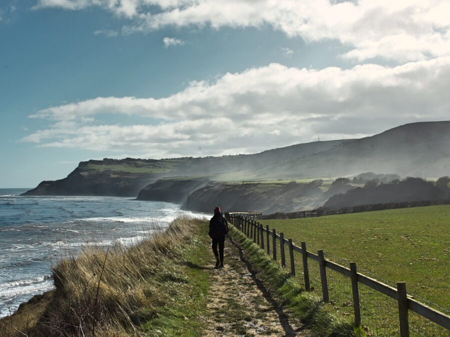 A man walking away from the camera along a grassy coast path with a red wooly hat on. There is a fence on his right with a large green field. On his left are grass bushes and the sea. In the distance is grassy hills and the sea air is creating a mist over them.