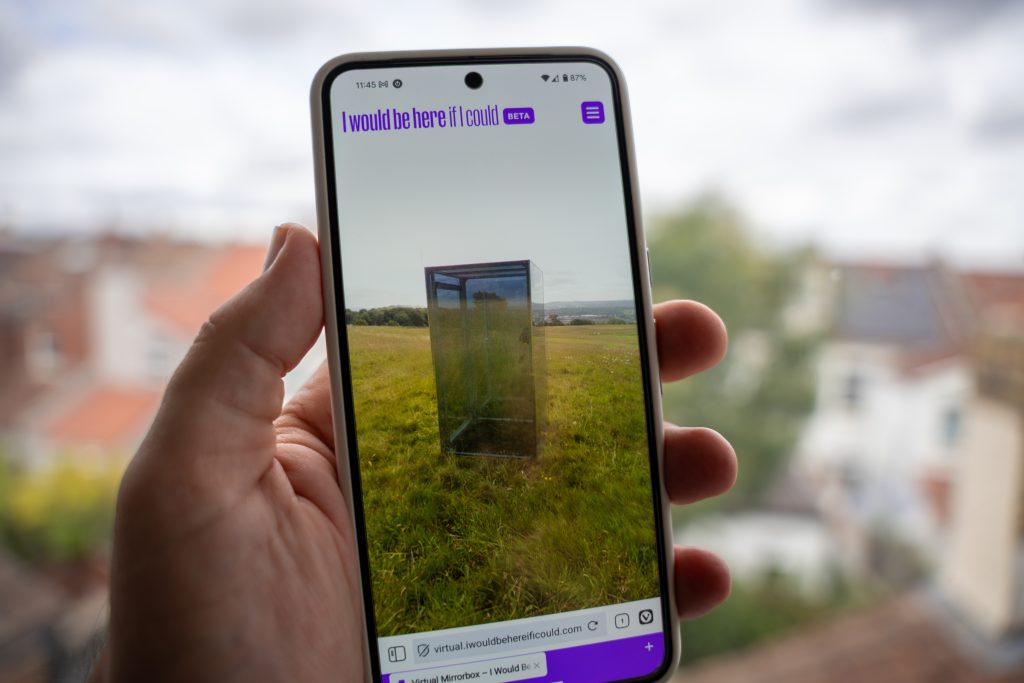 Photograph of a phone in a person's hand with the Virtual Mirrorbox experience on-screen.